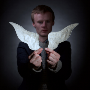 A man stands in the shadows with his hands held out in front of him holding two white paper wings