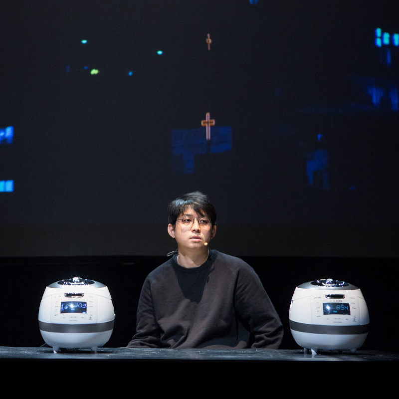 A man sits on stage at a desk with two rice cookers on either side