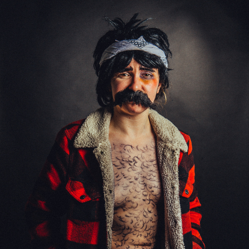 Natalie Palamides stands in a tartan jacket, wig and moustache with hair drawn on their chest