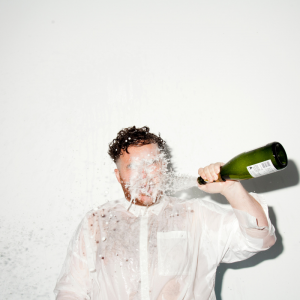 Scottee stands in a white shirt against a white background as he pours a bottle of champagne on himself