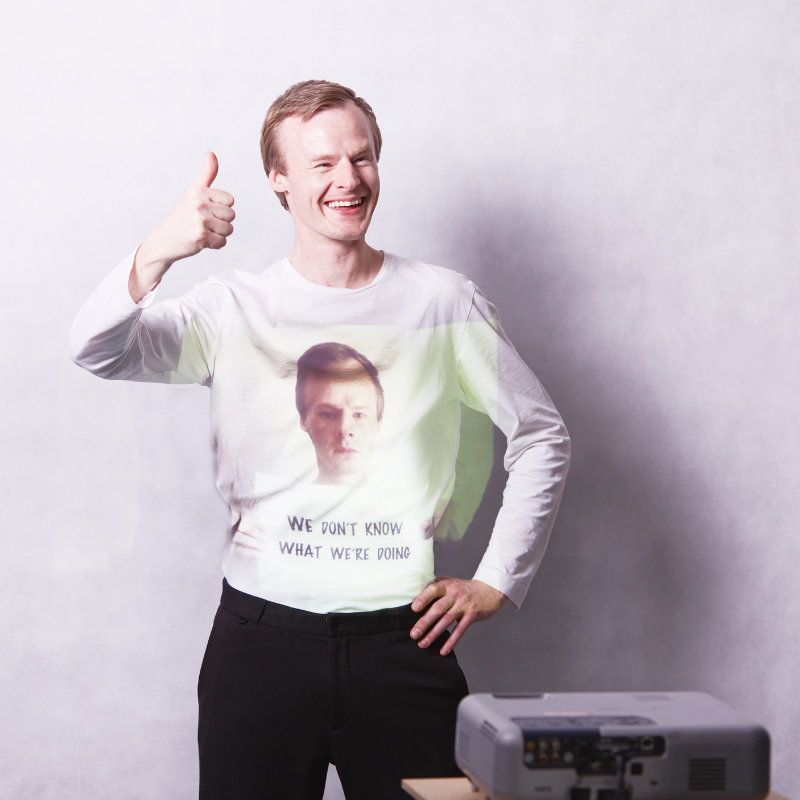 A man stands smiling with his thumb up, a projector projects the words 'we don't know what we're doing' onto his white jumper