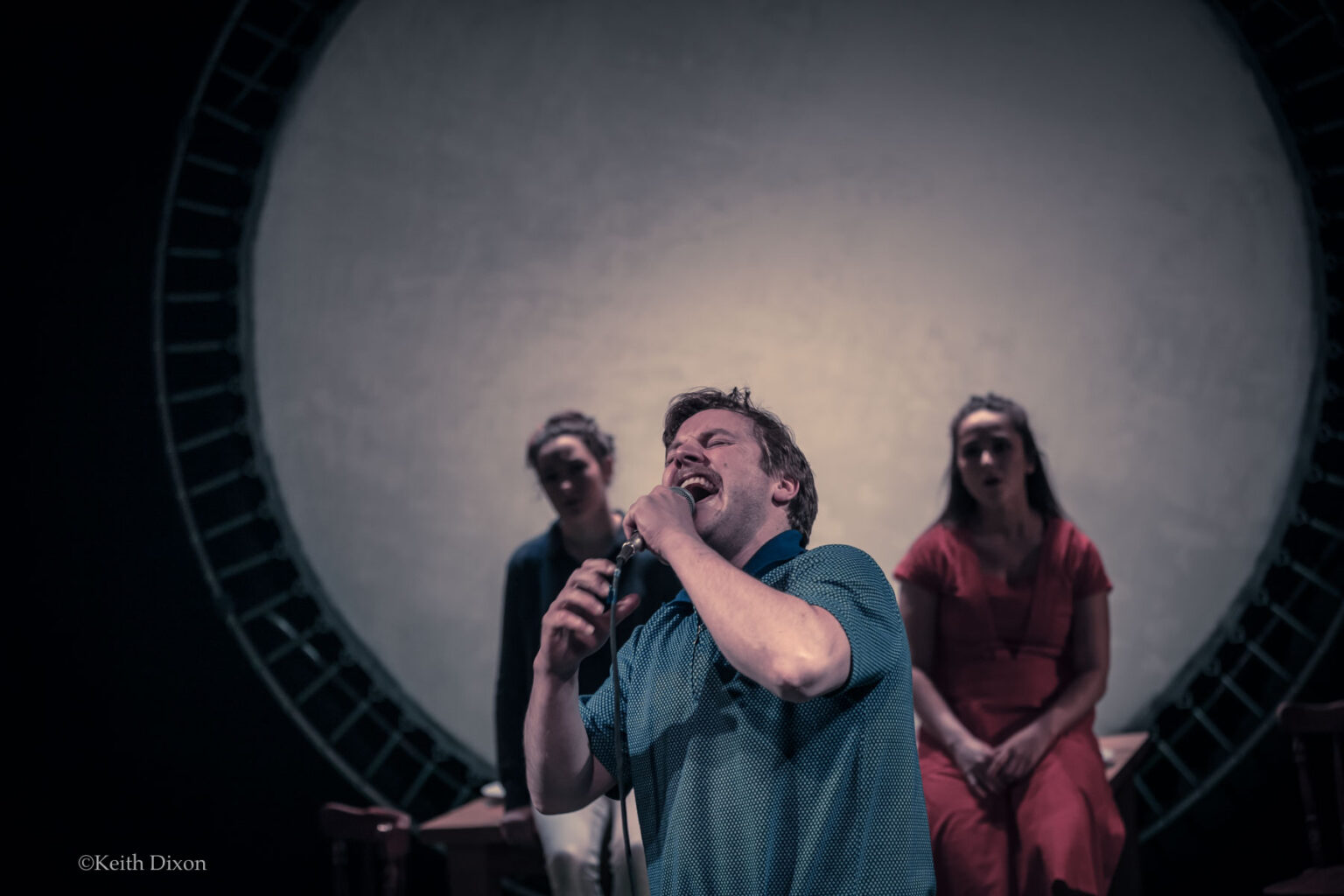 Two woman sit in front of a trampoline flanking a man in the middle singing passionately into a microphone.