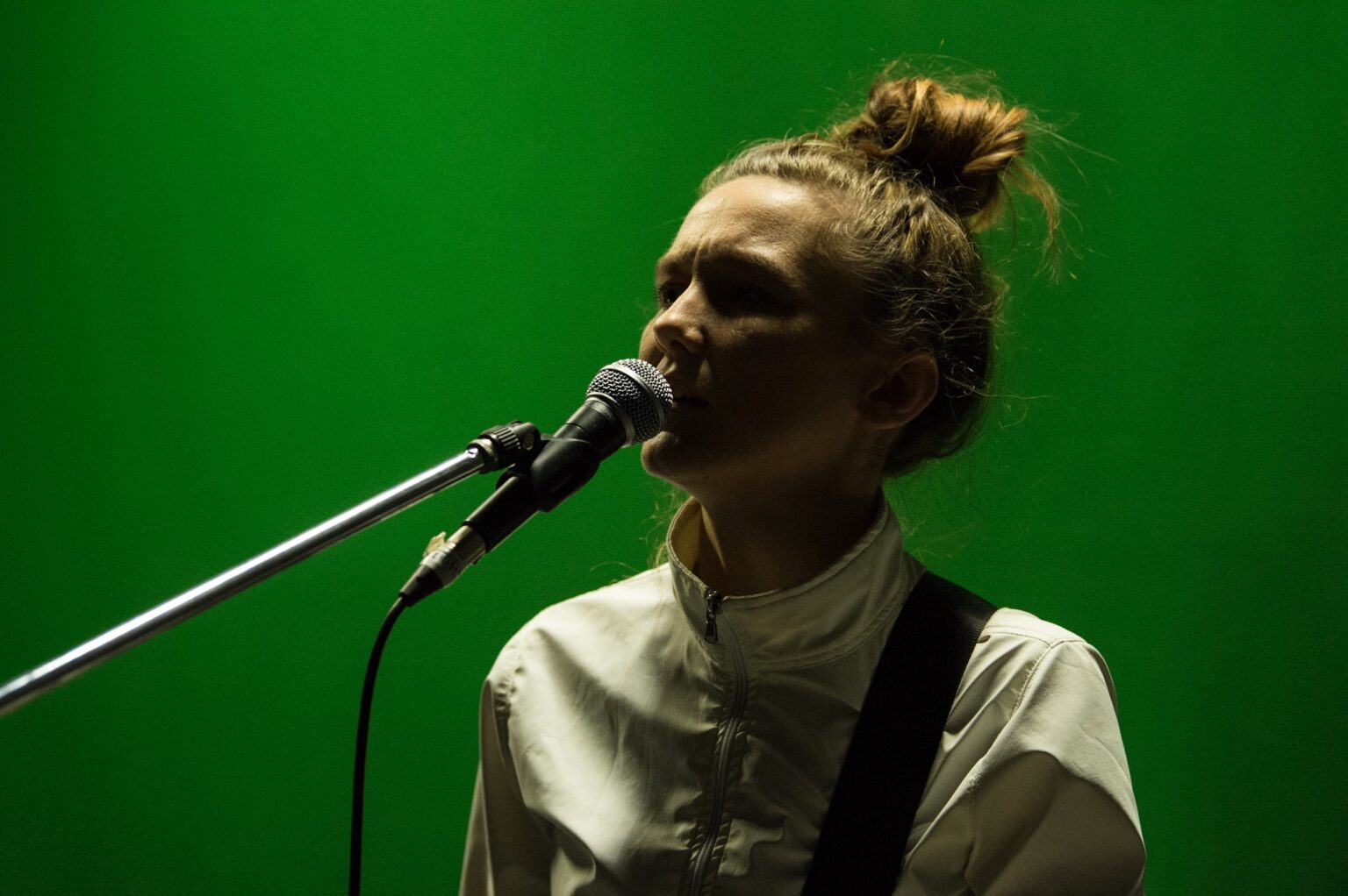 A woman with a bun in her hair wearing a white tracksuit standing in front of a green background holding a guitar and singing into a mic.