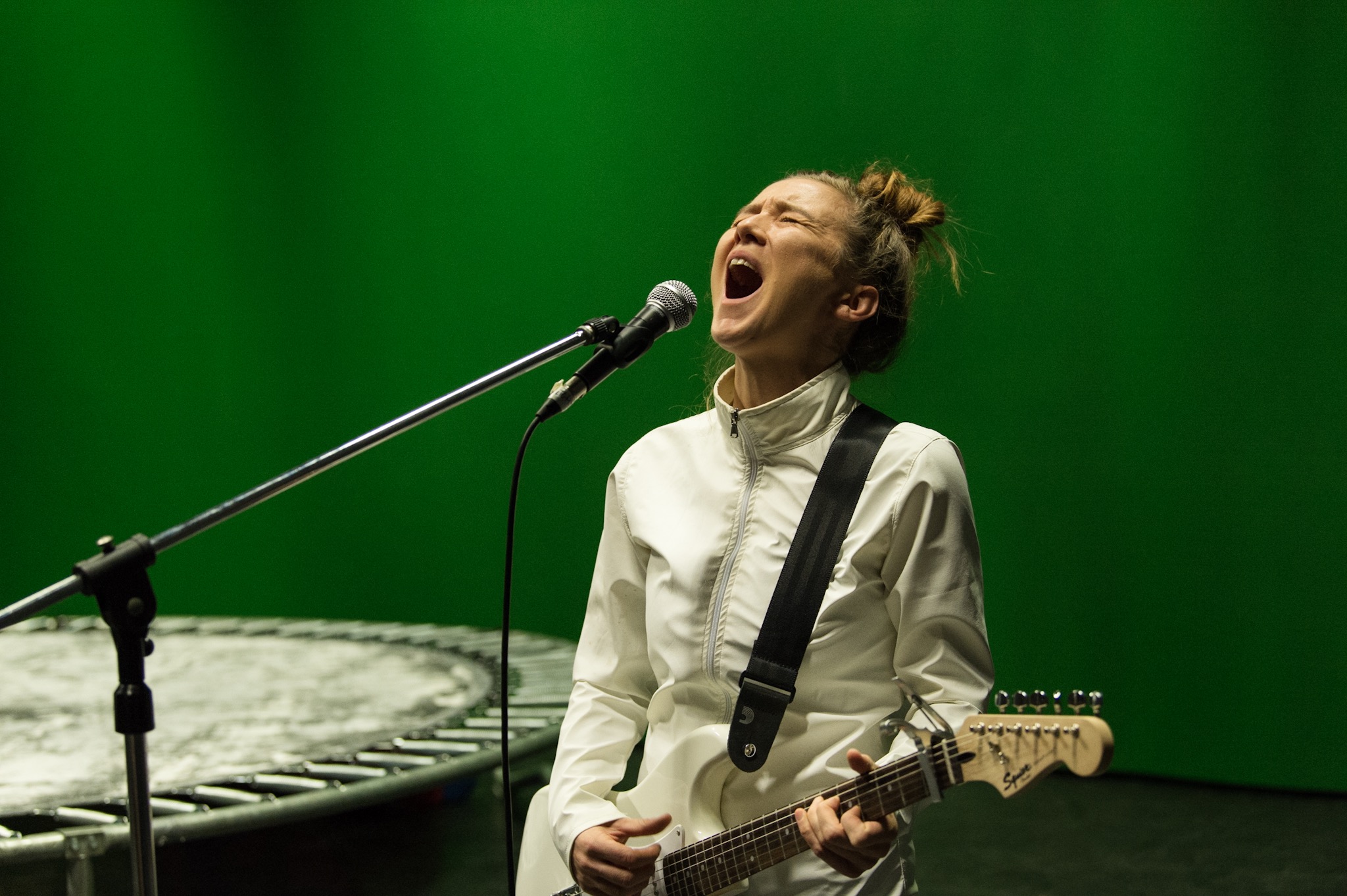 A woman with a bun in her hair wearing a white tracksuit standing in front of a green background holding a guitar and singing into a mic.