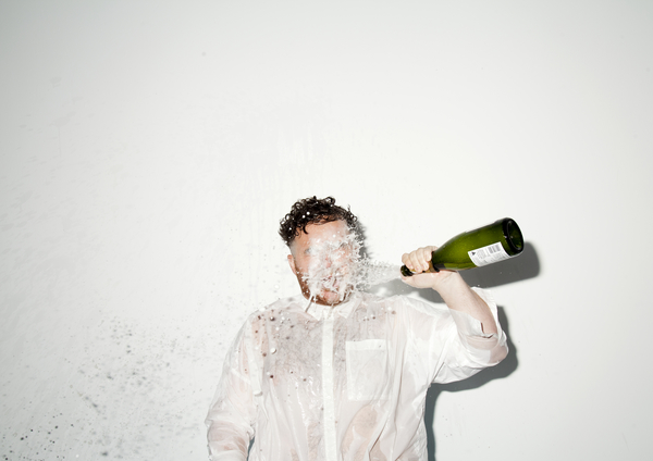 Scottee stands in a white shirt against a white background as he pours a bottle of champagne on himself