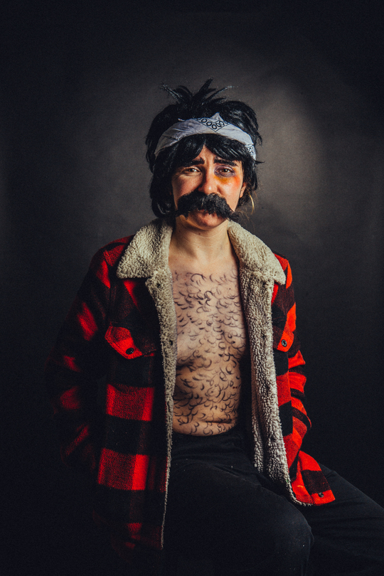 Natalie/Nate stands in a tartan jacket, wig and moustache with hair drawn on their chest