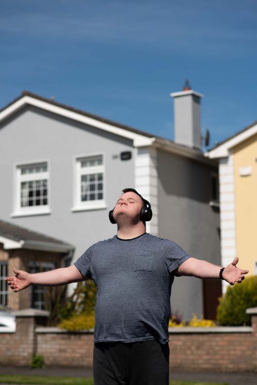A man wearing headphones stands in a suburban estate with his arms outstretched