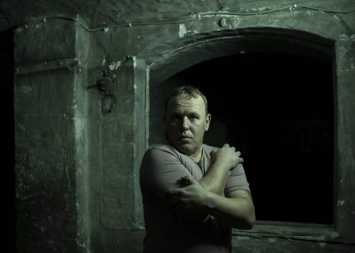 A man stands holding himself in a darkened prison cell