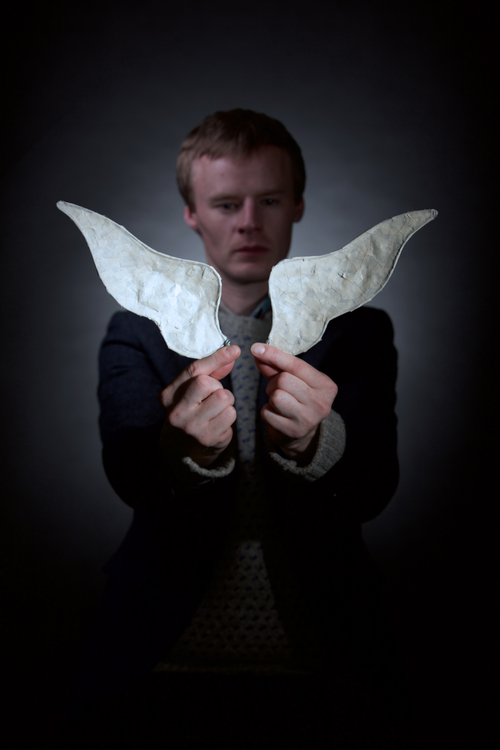 A man stands in the shadows with his hands held out in front of him holding two white paper wings