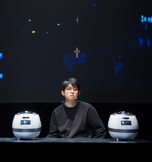A man sits on stage at a desk with two rice cookers on either side