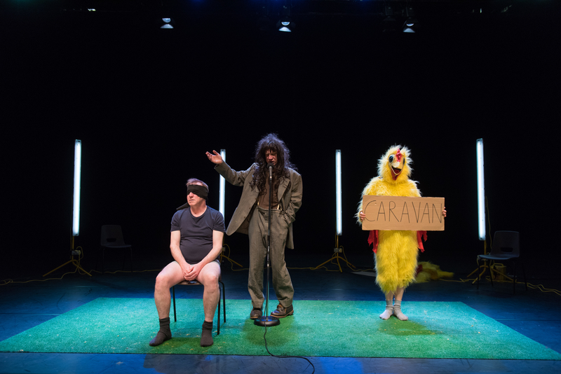 Show Image; Real Magic, conceived of and devised by Forced Entertainment company members, Directed by Tim Etchells presented at Dublin Theatre Festival. 3 characters in a sparse stage space; a blindfolded man without trousers sits on a chair, a dishevelled man in a grey suit and long brown wig gestures & speaks into a microphone in the centre. To the right of him a person in a chicken suit holds a cardboard sign saying Caravan