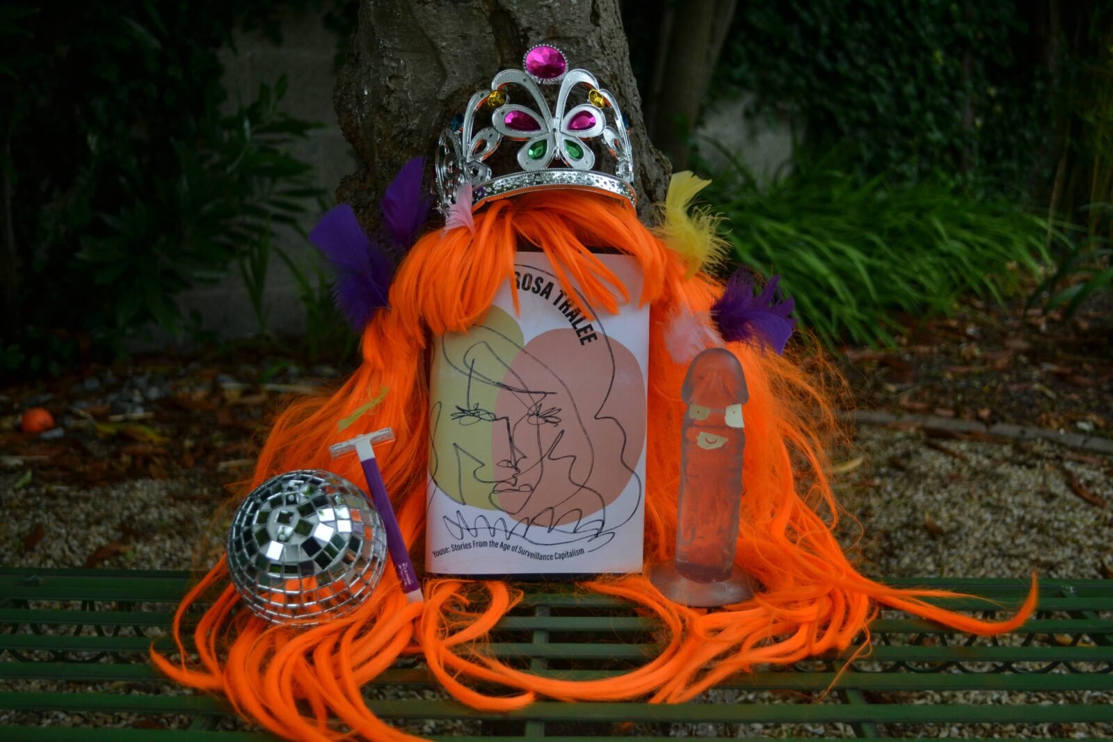 A long, bright orange wig in a plastic tiara with feathers, rests on a copy of Rosa Tralee's book with a mini disco ball and razer on the left and a sex toy on the other