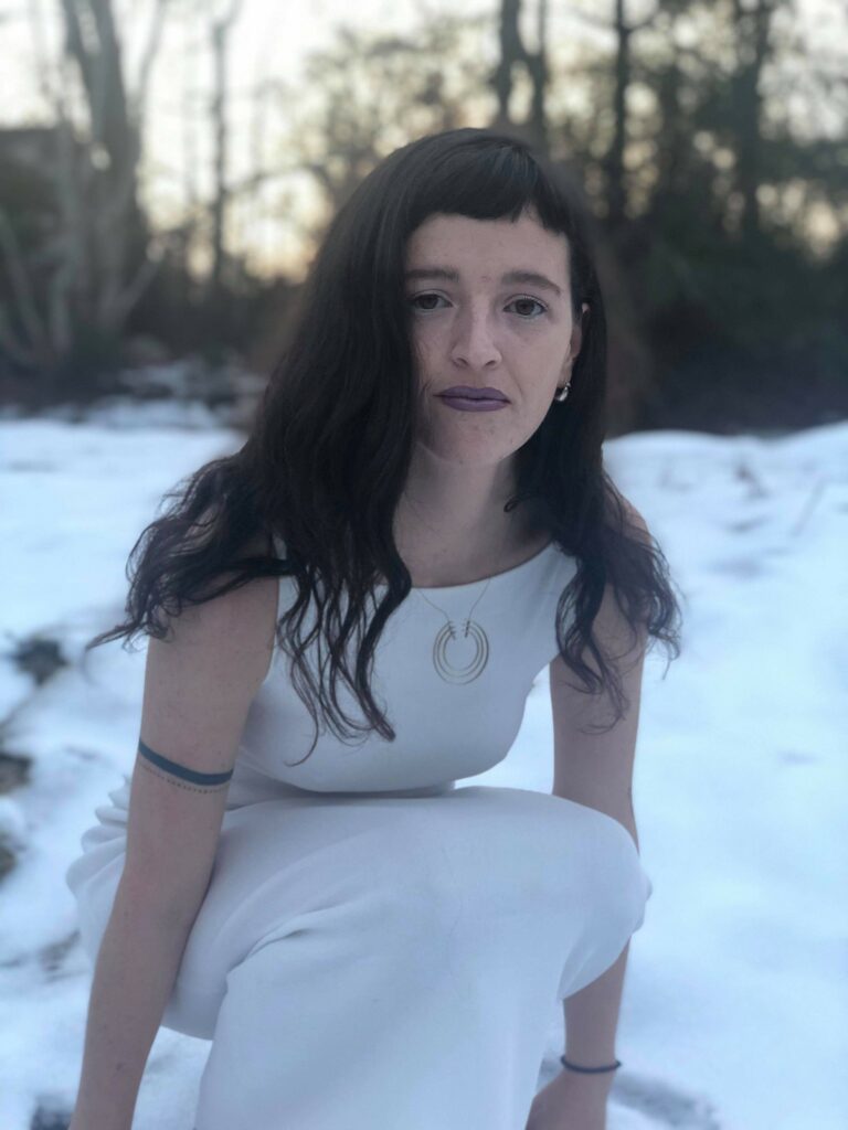 Photo of Aoife DeMille crouching in a white sleeveless dress and gold circular necklace in the snow with a wooded background 