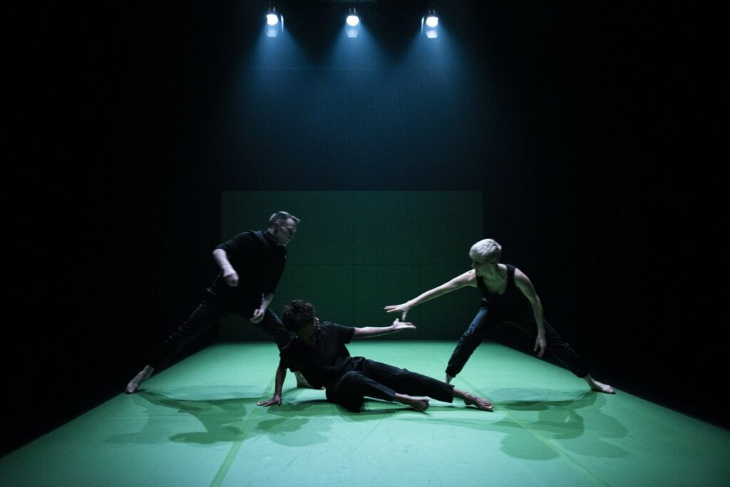 Three dancers dressed in black are positioned with a black wall behind them. They are illuminated by overhead lights and their bodies cast shadows on a bright green floor.