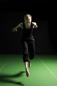 A white dancer with blonde hair is leaping off the floor, their body facing the camera. Both arms are stretched out behind them and their feet are off the floor, mid-jump. They’re wearing black trousers and a black sleeveless top. The room is dark with light illuminating them against a bright green floor with a black wall behind them.
