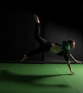 A dancer is positioned mid-leap, they are lunging forward with their positioned body at a horizontal angle, parallel to the floor. Both arms stretched out towards the floor. One leg is stretched up towards the ceiling, while the other one falls towards the floor. They are wearing black trousers and a green top. The room is dark with light illuminating them against a bright green floor with a black wall behind them.
