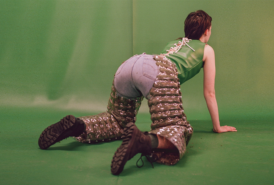 a photograph of a white person with a dark brown mullet is positioned on their hands and knees. They are facing their back towards you as they look away from the camera. They emphasise their ass by wearing a pair of PVC chaps which have been stuffed with green moss and pierced with metal eyelets. Under the chaps, they are wearing a pair of washed denim jeans. They are positioned in front of an infinitive wall of bright green leather. Their top is also made from this material and is held together using a corset technique.