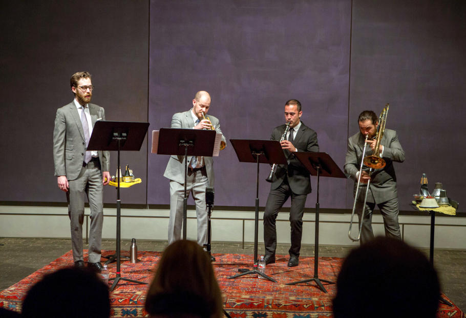 Four musicians wearing grey suits are positioned side by side, each in front of a music stand. They are playing wind instruments. They are standing on a red carpet and the wall behind them is made up of three large, dark grey panels. Audience heads form a silouhette of audience in the foreground of the image.