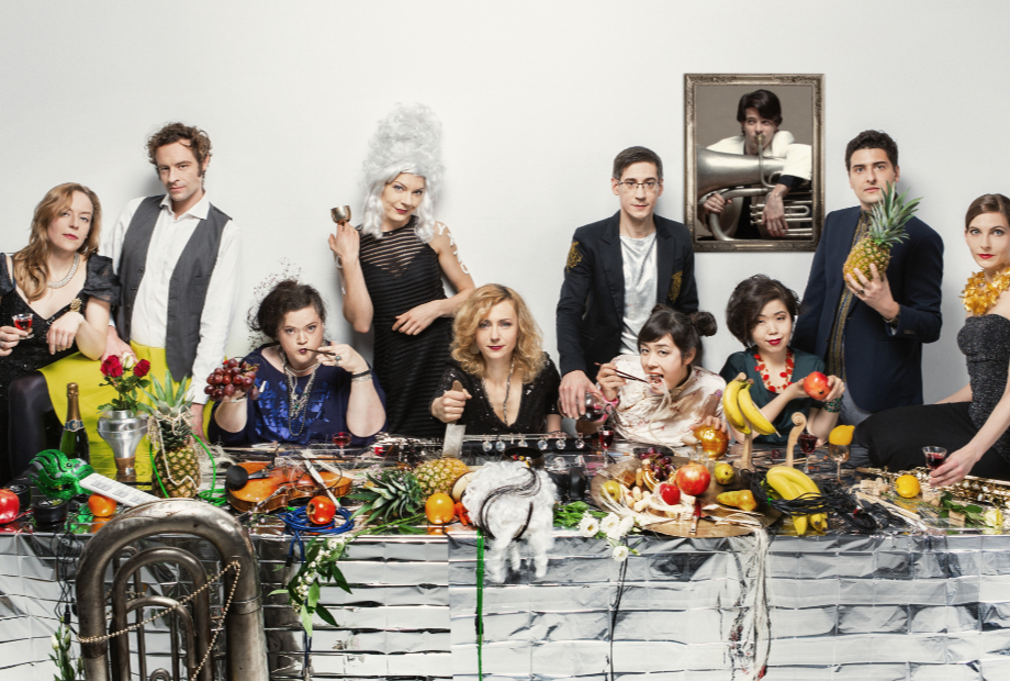 Ten members of the contemporary music ensemble, Ensemble Garage gather behind a long table. Four of the members are seated at the table and the remaining members stand behind. They are dressed formally. The table is covered in a silver table-cloth on top of which there is a lavish arrangement of food, wine, flowers and musical instruments. Some of the members of the ensemble are holding glasses and fruit. They are all looking at the camera. On the white wall behind the group, there is a portrait of a person playing a large brass instrument.
