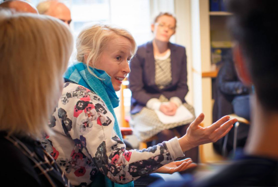 A group of people are sitting in a room engaged in discussion. The person in the centre of the room is in focus. The persons hands are raised and they are gesturing and looking at the other participants.