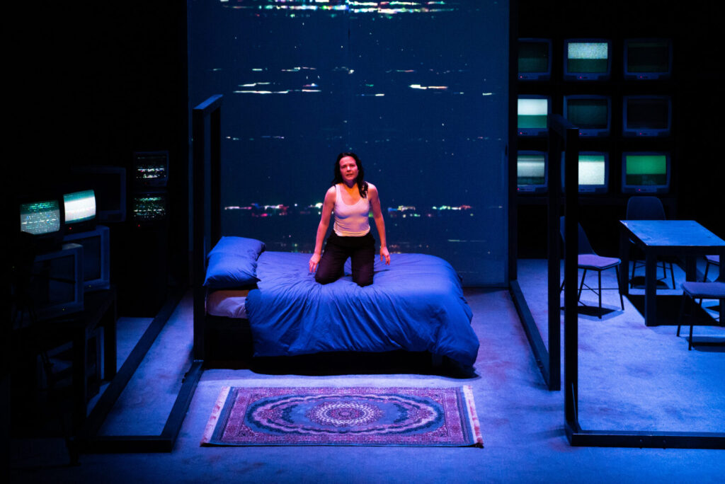 A white woman with long dark hair wearing black trousers and a white vest kneels upright on a double bed with blue sheets. Her expression is serious, her arms hang loosely to her sides. The floor is grey carpet, with a persian rug spread out in the foreground in front of the bed. On the back wall, TV static is projected from floor to ceiling. To the left of the image, multiple televisions of varying sizes are stacked on top of each other. Some are off and some are displaying static.