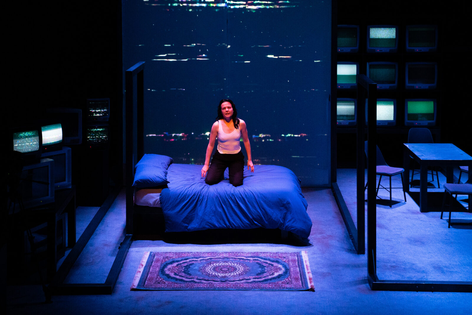 A white woman with long dark hair wearing black trousers and a white vest kneels upright on a double bed with blue sheets. Her expression is serious, her arms hang loosely to her sides. The floor is grey carpet, with a persian rug spread out in the foreground in front of the bed. On the back wall, TV static is projected from floor to ceiling. To the left of the image, multiple televisions of varying sizes are stacked on top of each other. Some are off and some are displaying static.