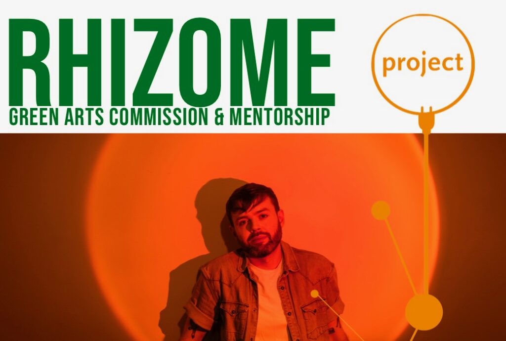 A graphic with text and a photograph. The banner at the top of the graphic contains text that reads :RHIZOME: GREEN ARTS COMMISSION & MENTORSHIP", Project's Logo is positioned beside the text, and is outlined in orange. An orange plug and series of abstract shapes connect the banner to the photo below. In the photo, artist Louis Haugh stands facing the camera. He has brown hair and a short beard. He is wearing an unbuttoned shirt with a white t-shirt underneath. He is standing in an orange, glowing spotlight that forms a circle behind him.