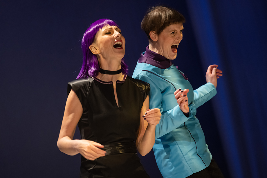 Two cast members stand side by side with their shoulders touching. One cast member has long purple hair, and is wearing a black, short sleeved top and a black neckband. The other cast member has short, brown hair and is wearing a long-sleeved, light blue top with a purple collar. Their expressions are animated and their mouths are wide open, mid laugh, or shout. The background is dark blue.