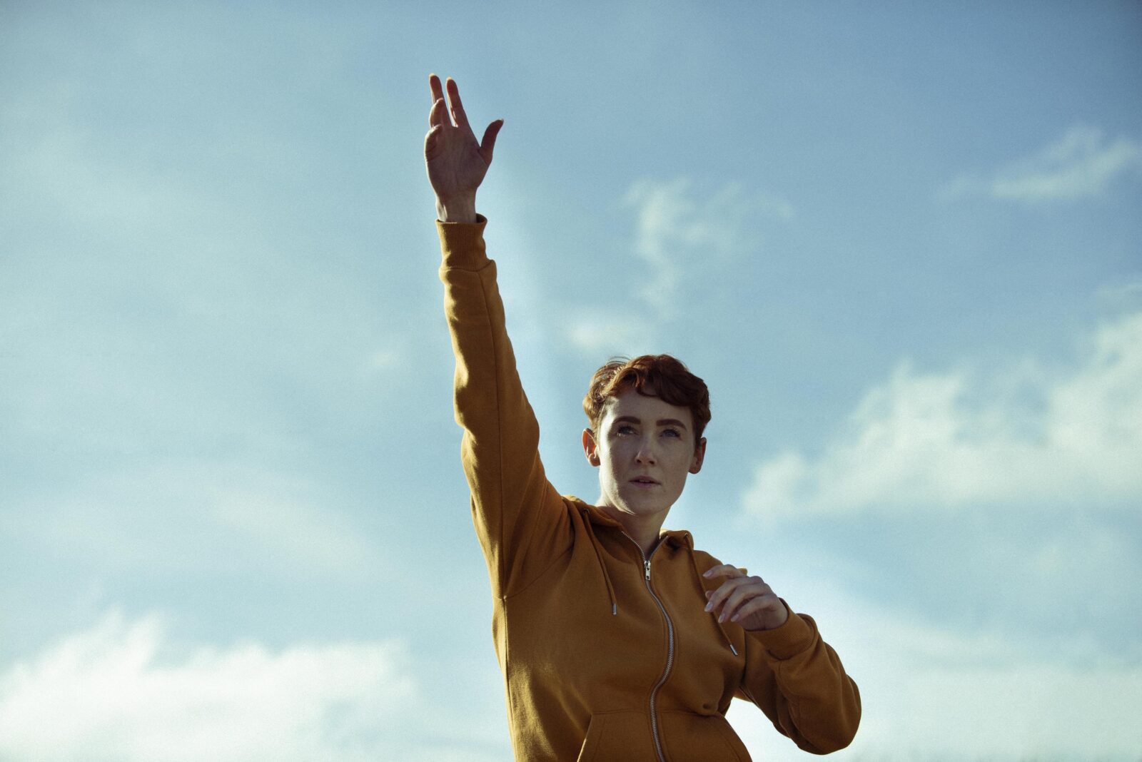 A person poses in the centre of the image. In the background there is blue sky with a few scattered white clouds. The person has short hair, and is wearing an orange zipped top. One arm points towards the sky and they hold the other arm at an angle across their chest.