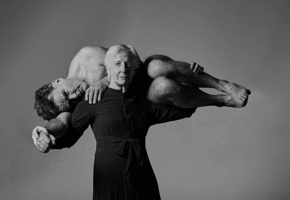 A black and white image. A woman stands in the centre of the image, she is wearing a black dress. She holds a man on her shoulders. He is unclothed and is wrapped around her neck and shoulders. The background of the image is grey