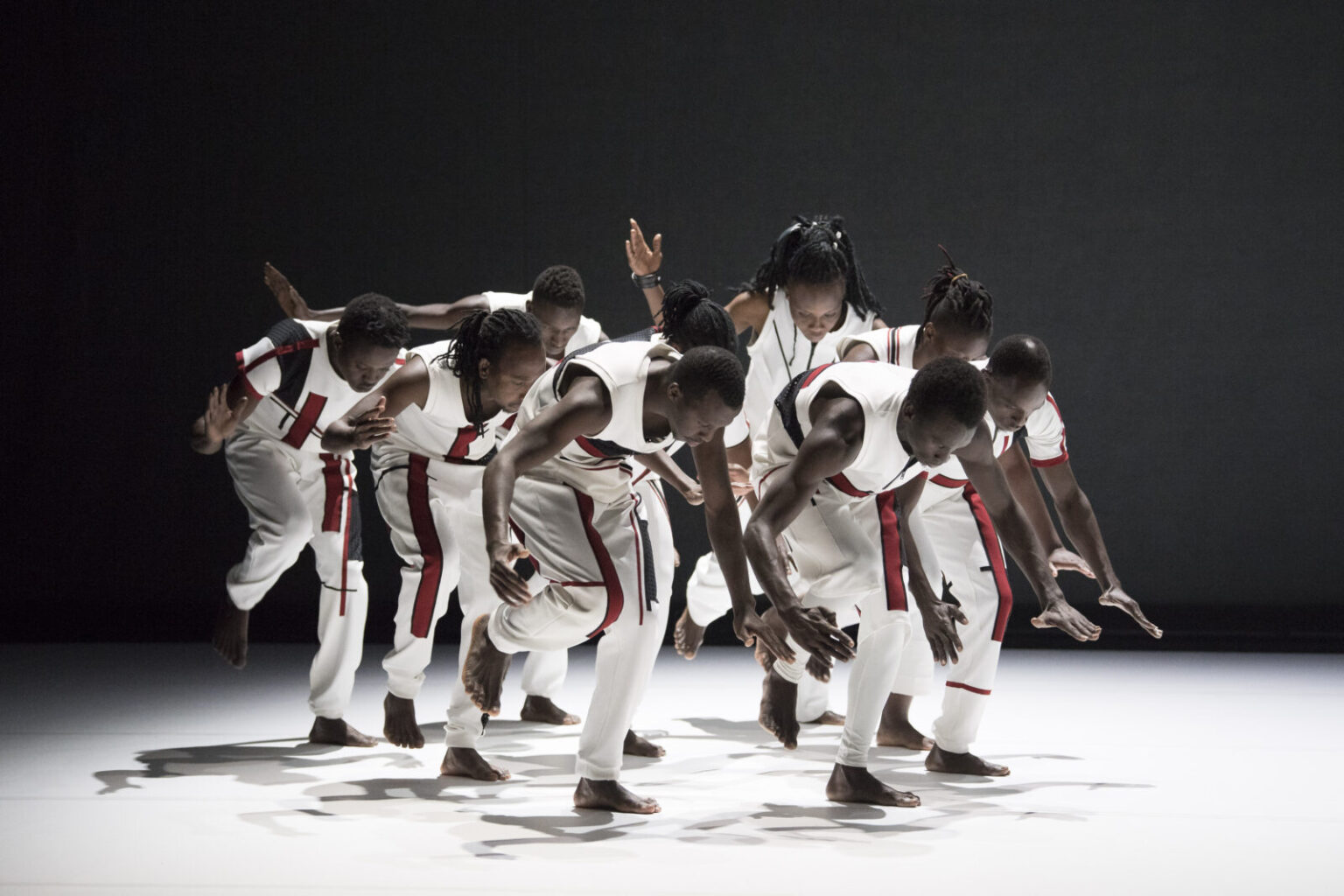 A group of African dancers move in unison across a white floor. They are each standing on one foot, and are looking and leaning towards the floor, with their arms outstretched. The dancers are wearing white clothing, with a red and black graphic detail printed on the trousers and short sleeved tops. The background of the image is black