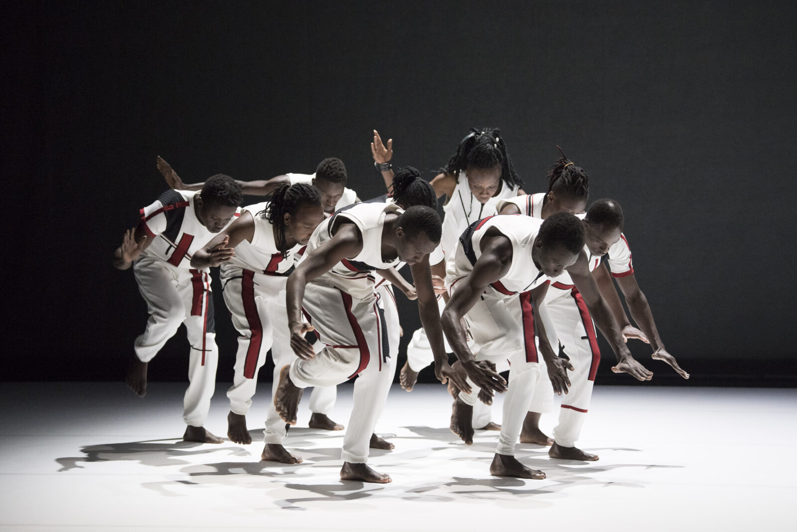 A group of African dancers move in unison across a white floor. They are each standing on one foot, and are looking and leaning towards the floor, with their arms outstretched. The dancers are wearing white clothing, with a red and black graphic detail printed on the trousers and short sleeved tops. The background of the image is black