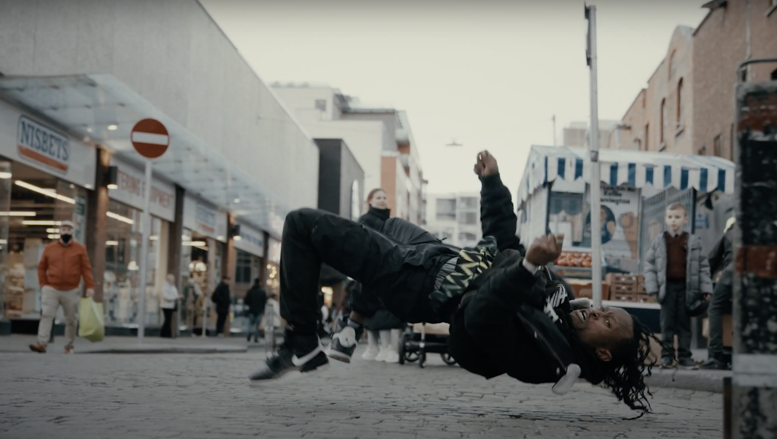 Image Description: An image of Tobi Omoteso dancing on Moore Street. Tobi is in the foreground of the image, his body is arched and lifted off the cobble stones on the ground below, he is in motion and looking towards the camera. There are people in the background of the image and a young person in a school uniform looks at him. There are shops and a fruit and veg stall in the background. The sky is grey. The image is a still, taken from a film shot by Jacek Snochowski.