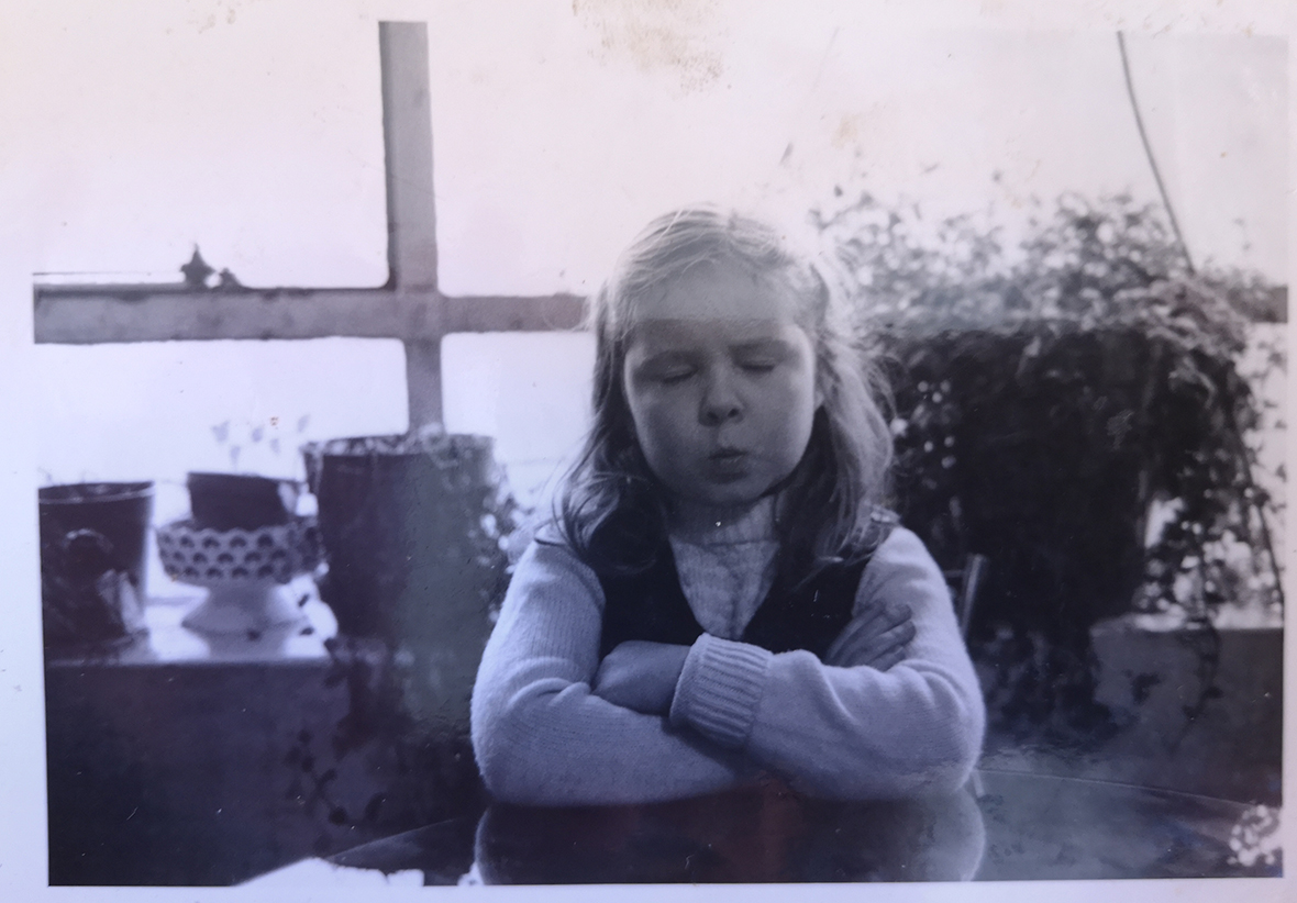A photograph of Veronica Dyas as a young child. She sits in the centre of the image facing the camera at a table with her arms crossed and her eyes closed. Her lips are pursed. Behind her is a window cill, with bowls and potted plants lining it. The photograph is black and white. Taken by Pat McGrath, circa 1987.