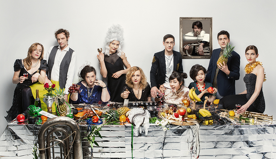 Image Description: Ten members of the contemporary music ensemble, Ensemble Garage gather behind a long table. Four of the members are seated at the table and the remaining members stand behind. They are dressed formally. The table is covered in a silver table-cloth on top of which there is a lavish arrangement of food, wine, flowers and musical instruments. Some of the members of the ensemble are holding glasses and fruit. They are all looking at the camera. On the white wall behind the group, there is a portrait of a person playing a large brass instrument.