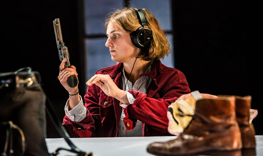 A white woman with loose, short hair sits at a table on which the camera is positioned. In the foreground on the table is a pair of brown leather boots and a small black backpack. The woman is wearing a red jacket over a shirt and a large pair of headphones. She is holding a gun, pointing towards the ceiling, which she is looking at. There is a window behind her.