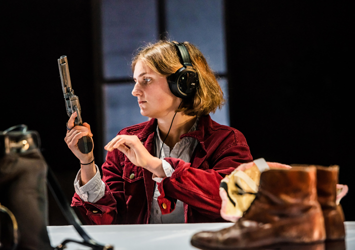 A white woman with loose, short hair sits at a table on which the camera is positioned. In the foreground on the table is a pair of brown leather boots and a small black backpack. The woman is wearing a red jacket over a shirt and a large pair of headphones. She is holding a gun, pointing towards the ceiling, which she is looking at. There is a window behind her.