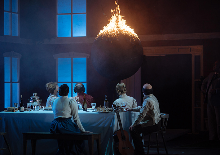 A group of 3 women and 2 men people sit with their backs to the camera around a table, as a man in a suit stands in the shadows to the right. They are white and their costume and hair is Victorian in style. The table is covered with a white tablecloth and on it sit bottles of wine, crystal glasses, a silver ice bucket, a straw hat and a game of chess. One of the seated men has a guitar propped up next to him. All of the people are looking at a large black wrecking ball hanging down from the ceiling, the top of which is on fire. The windows in the room are letting in dusky light and there is a large arched doorway open at the back.