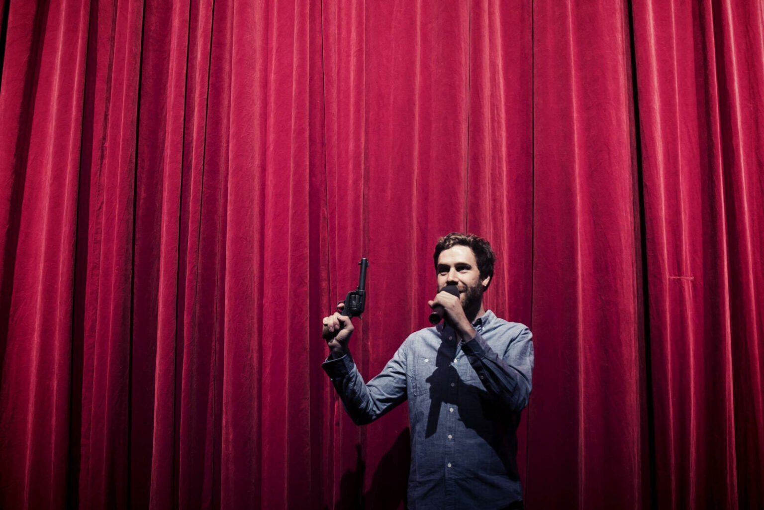 A white man with a beard wearing a shirt stands in front of a red theatre curtain. He's visible from the waist up. He is holding up a gun in one hand that points towards the ceiling. In the other hand, he's holding a microphone up to his mouth.