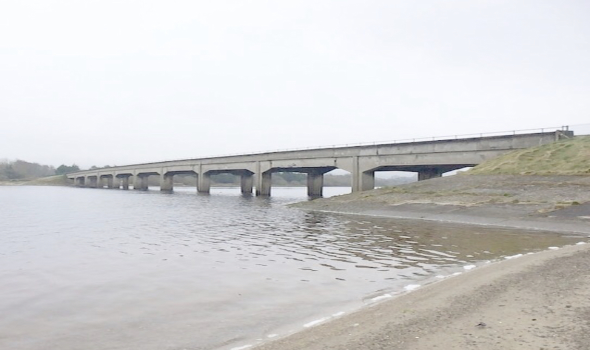 A brutalist concrete bridge spans a river. The colours of river, bridge, and sky are greys and browns.