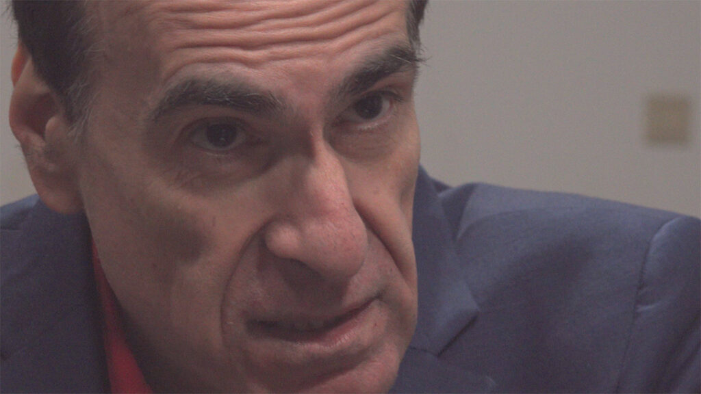 A still from the film Rope; shows a close-up of a man's face. He wears a muted blue suit-jacket and red shirt. He has dark hair and strong features and his face is craggy and lined. His head is tilted down and his eyebrows are up in an expression of scepticism or questioning.