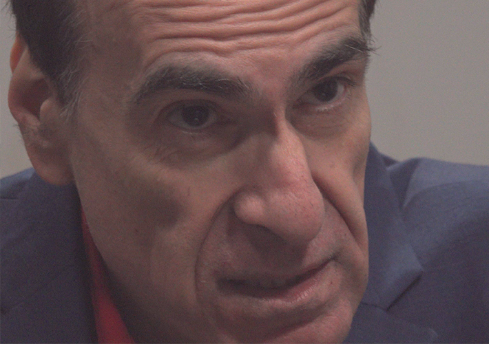 A still from the film Rope' shows a close-up of a man's face. He wears a muted blue suit-jacket and red shirt. He has dark hair and strong features and his face is craggy and lined. His head is tilted down and his eyebrows are up in an expression of scepticism or questioning.