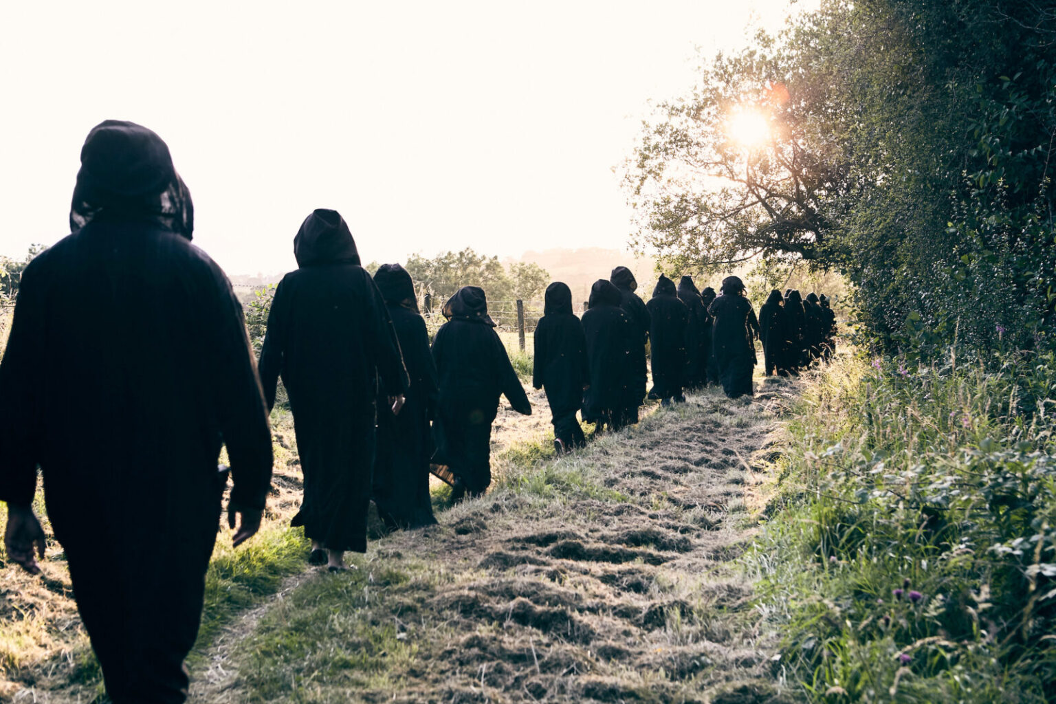 A landscape photograph of a long line of people in black hooded robes walk along a path at the edge of a field with their backs to the camera.