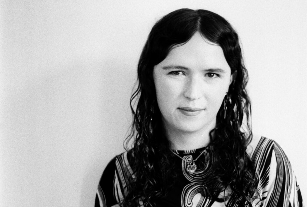 A black and white photo of Iarlaith Ni Fheorais. Iarlaith is standing in front of a white wall, to the right of the frame. Iarlaith is a white woman, with long curly brown hair. She is wearing a tight ruched top with a spirally print with large sleeves, a pendant necklace and long earrings. She has a slight smile.