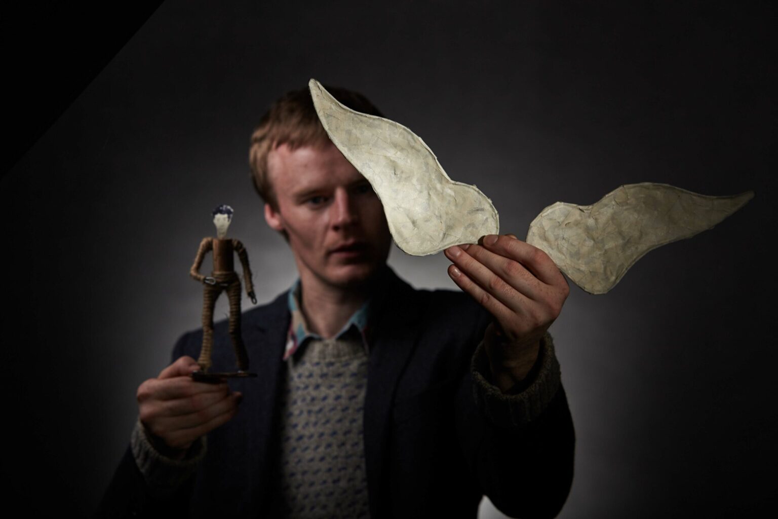 A young man with blonde hair dressed in a shabby suit faces us is holding a small pair of toy wings in his left hand and a small hand-made figure of a man in his right hand.