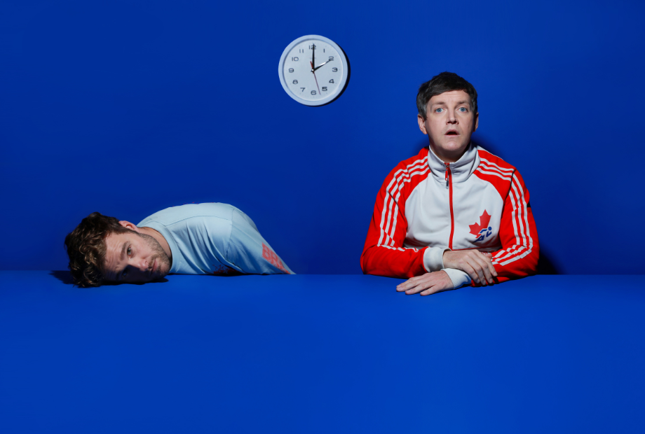 Two men (30s) seated in front of a clock, one with his head resting on a table. Foreground and background are a strong royal blue colour.