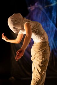 A portrait photo of Ali Clarke, performing her piece Multitudes; Future Memories. Ali has a pale complexion and is wearing beige trousers and a short-sleeve white top with thin black stripes. She has beige fabric wrapped around her head, with just the shape of her face pushing through the fabric. She is standing with her right side to the camera. Her back is arched with her face and chest directed towards the ceiling. Her arms are reaching out to either side behind her. She stands in a dark space, with an unidentifiable object in the background which is lit in blue and purple.