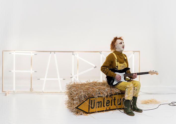 A person sits down holding a guitar. They are wearing an abstract mask. They wear yellow dungarees and green welly boots, sitting on a bale of hay. The background is white and a black and white object hangs above.