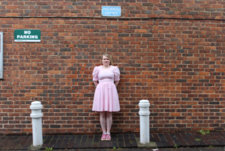 A juvenile looking woman in a pink party dress stands in front a high brick wall with a ‘No Ball Games’ sign.