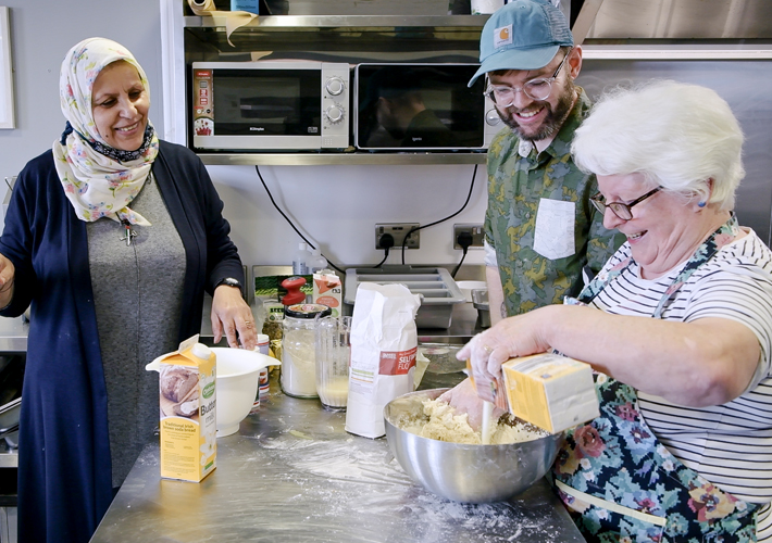 There are three people in the image. They are all smiling. They are making bread at a kitchen counter. There is a woman standing to the left wearing a black headscarf and a black dress. There is a man standing to the right of the counter wearing a baseball cap and glasses. He has short brown hair. There is a woman on the right in the foreground wearing an apron and a white t-shirt. She has short white hair and is also wearing glasses. She is pouring butter milk into a big mixing bowl. There are other ingredients on the counter in front of them.